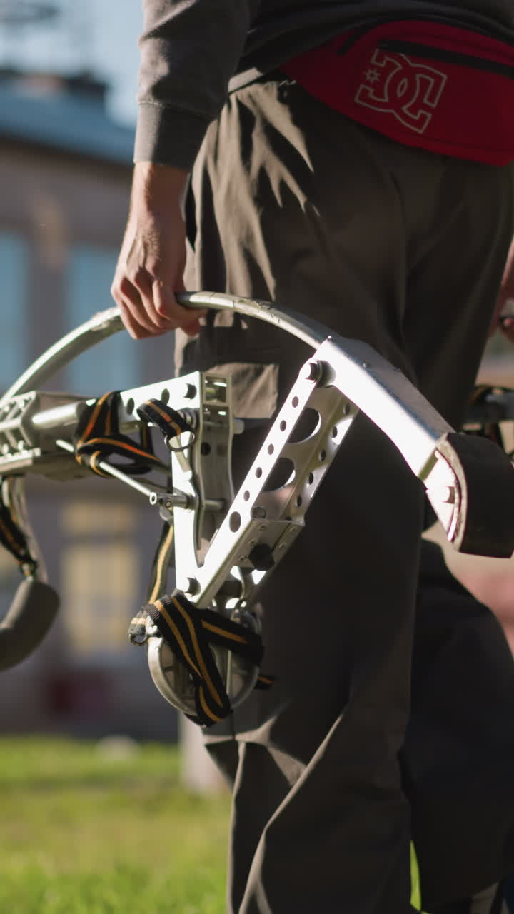 검은색 바지를 입고, 어두운 회색 장갑 셔츠를 입고, 눈에 보이는 색 로고가 있는 빨간색  가방을 들고 있는 남자. 배경은 잔디과 멀리 있는 건물로 흐릿하다.