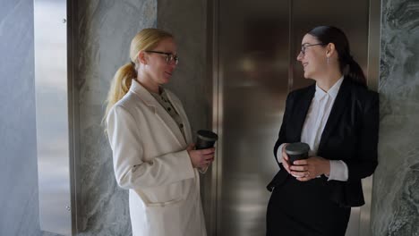 Couple-of-happy-businesswomen-in-a-white-and-black-business-suit-talking-while-drinking-coffee-during-a-break-from-work-in-a-modern-office