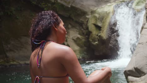 Mädchen-Mit-Lockigem-Haar-Im-Bikini-Sitzt-Auf-Den-Felsen-Mit-Einem-Fließenden-Wasserfall-Im-Hintergrund