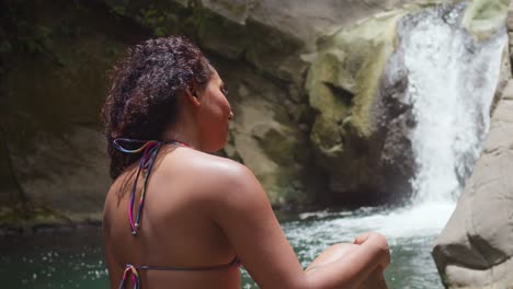 curly hair girl in bikini sits on the rocks with a flowing watefall in the background