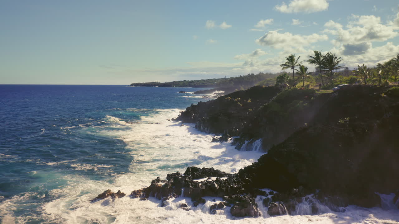 Powerful waves surge and crash against rugged volcanic cliffs under a bright tropical sky, the deep blue Pacific meeting the raw, untamed edges of an island paradise