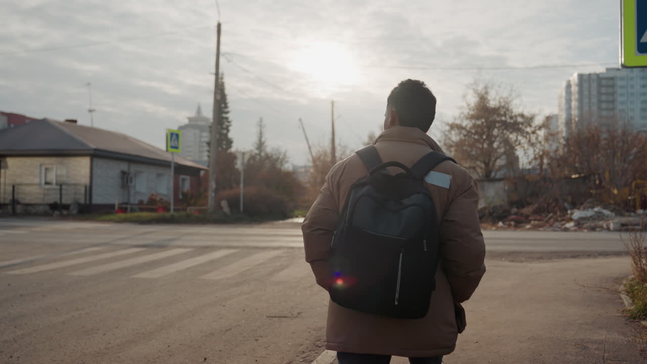 Un viajero con mochila, en un cruce de carretera por la mañana, inspecciona un neumático abandonado. El sol está bajo en el horizonte, se ven casas suburbanas y una calle tranquila. La silueta, vista de espaldas, revela su intención de reparar un pinchazo, con las manos buscando herramientas.