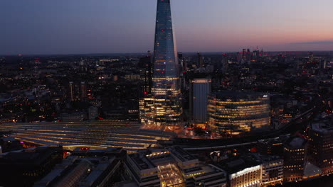 Diapositiva-Y-Toma-Panorámica-De-La-Estación-De-Tren-Del-Puente-De-Londres-Iluminada,-El-Edificio-De-Noticias-Y-El-Rascacielos-De-Fragmentos.-Vista-Aérea-Nocturna-De-La-Ciudad-Después-Del-Atardecer.-Londres,-Reino-Unido