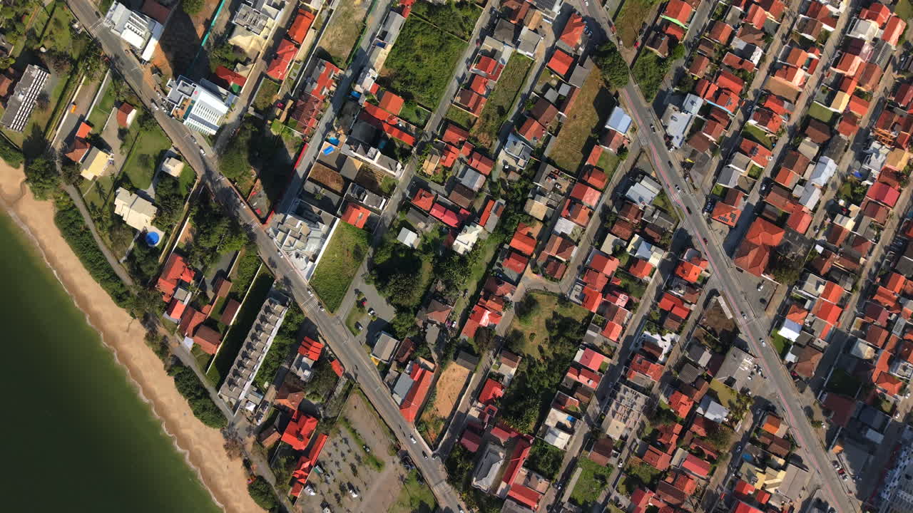 Overhead drone glides above sunny seaside neighborhood, showcasing vibrant orange clay rooftops and lush green yards