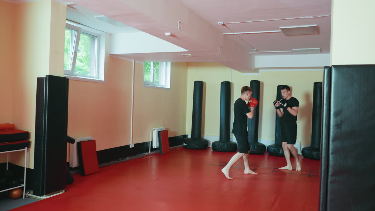 Practicantes de kárate combatiendo en un gimnasio de artes marciales, donde uno de ellos da una potente patada frontal mientras su oponente se defiende con guantes sobre una colchoneta roja, rodeados de sacos de boxeo e iluminados por la luz natural que entra por las ventanas.