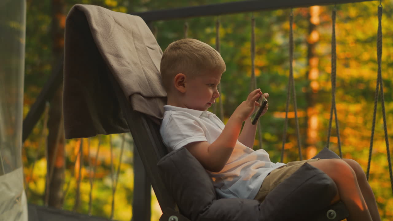 Little boy plays on contemporary smartphone enjoying pleasant evening. Amazing nature scenery surrounds toddler child sitting in comfortable armchair at sunset