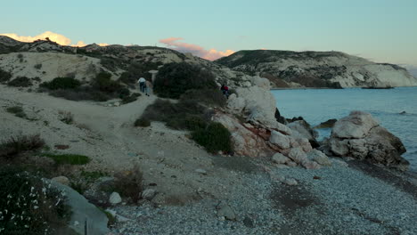 tourists explore aphrodite's rock at sunset, walking on rocky pebble beach in paphos, cyprus - crane down shot