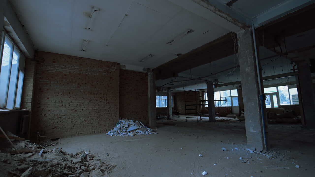 Entering the wide doorway in the brick wall with broken window. Spacious hall of a building with some construction debris left on the floor.