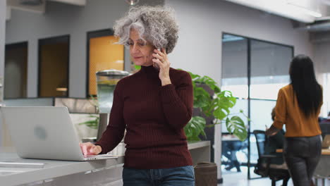 mature businesswoman on phone working on laptop in kitchen area of busy modern office