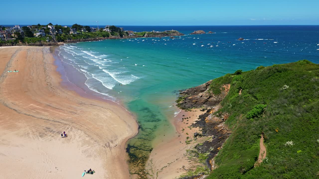 Turquoise water gently hitting sandy Saint-Lunaire Beach with lush green headland in drone view, Saint-Lunaire, Bretagne, France.