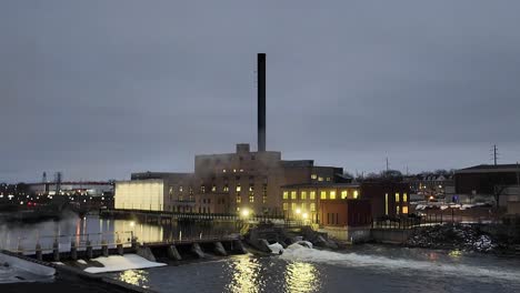 Frühmorgendliche-Aufnahme-Einer-Fabrik-Mit-Schornstein-Und-Damm-Auf-Der-Anderen-Seite-Des-Flusses-Am-Morgen