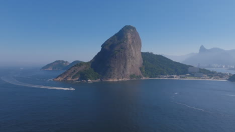 wide aerial footage of sugarloaf mountain with a boat crossing in front of it and rio de janiero in the background during the day