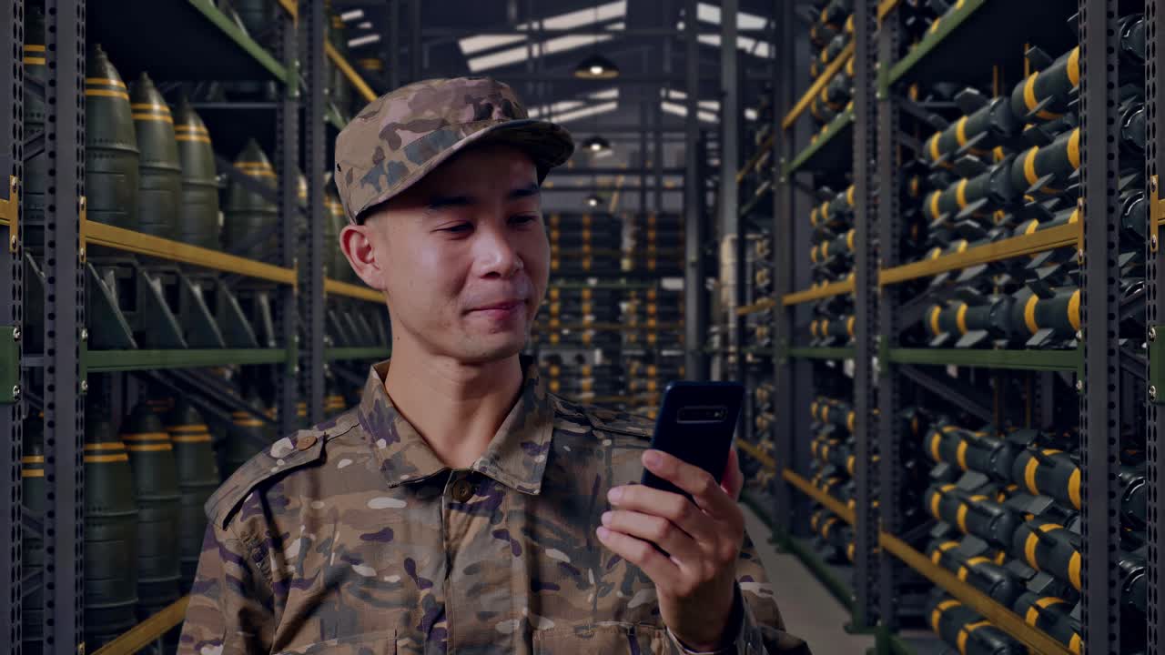 Primo piano di un militare asiatico che saluta con la mano durante una videochiamata su smartphone mentre si trova in un deposito di munizioni militari