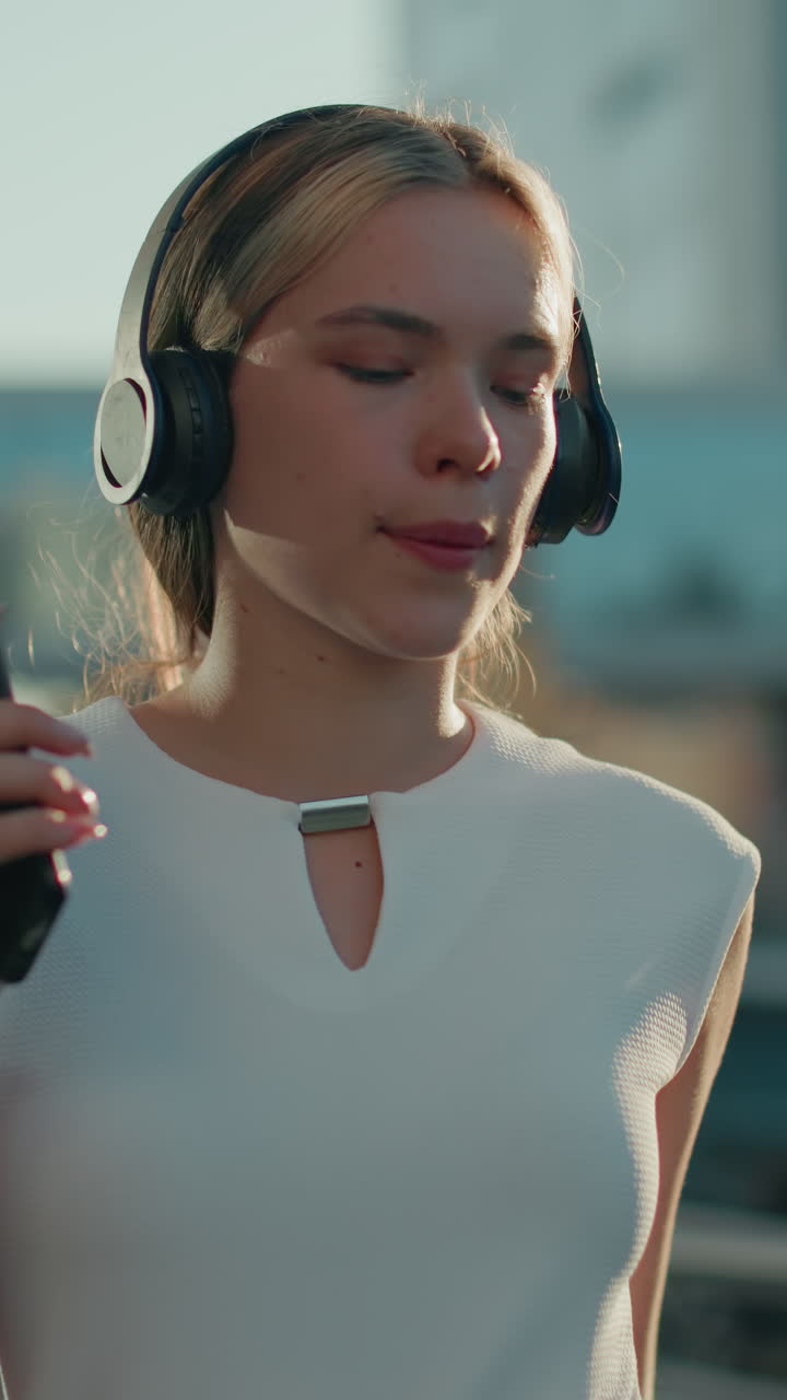 Golden sunlight ambiance surrounds young lady enjoying music with headphones and phone in hand while dancing gracefully in urban setting with slight blur background of cars and buildings