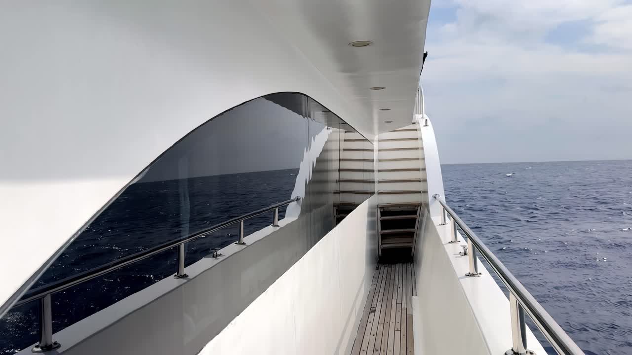 Eine elegante Luxusyacht gleitet an einem hellen Sommertag durch tiefblaue Ozeangewässer, mit einer Draufsicht auf einen schmalen Teakholz-Gang neben gewölbten getönten Fenstern unter einem klaren, sonnigen Himmel.