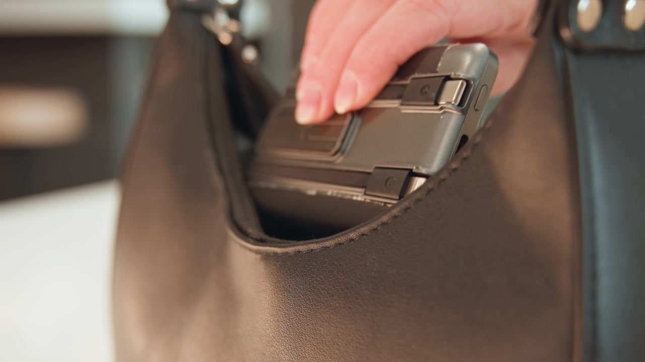 Vista ravvicinata di una mano che inserisce uno smartphone nella tasca anteriore di una borsa in pelle martellata marrone, con le dita che afferrano il dispositivo e il bordo in pelle che si piega mentre il telefono scivola all'interno, con luce soffusa e profondità di campo ridotta