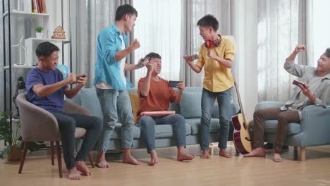 group of young asian friends with musical instruments celebrating winning game on smartphones while sitting on a sofa at home