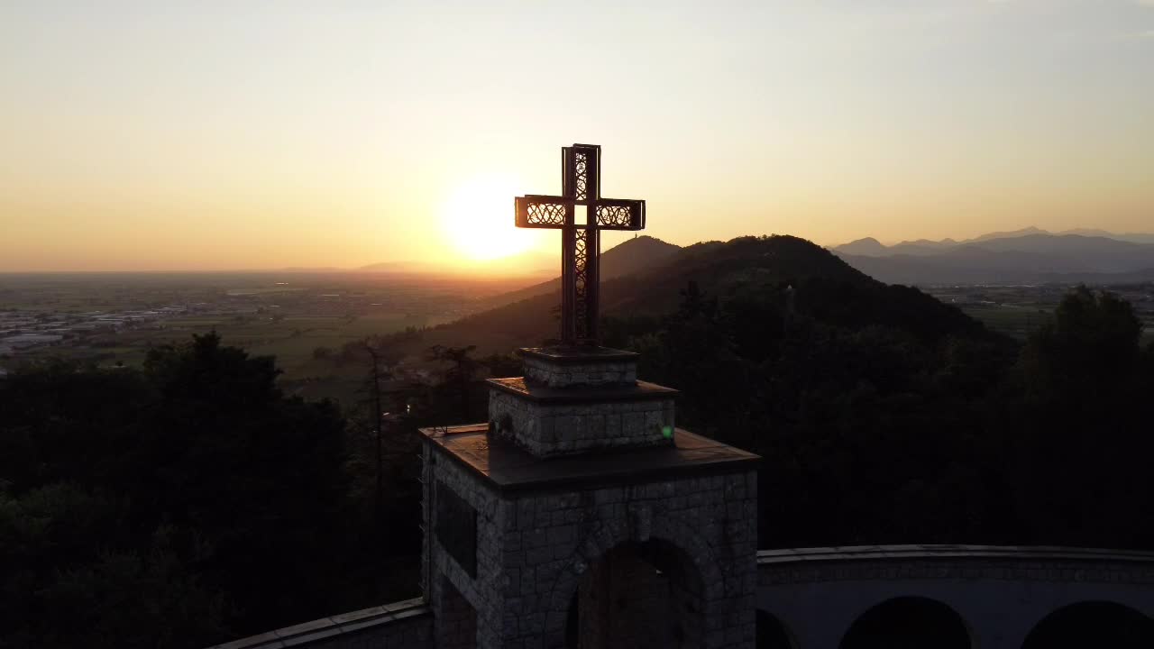 A stunning aerial shot of a sunset behind a large cross on a hill in Italy showcases the beauty of the landscape and the golden light.