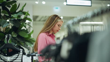 white lady in pink top browses through hanged clothes in shopping mall with green plants behind her, other shoppers are blurred in the background, also picking clothes from racks in a well lit mall