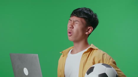 공을 들고 있는 아시아 소년이 녹색 스크린 스튜디오에서 노트북으로 축구를 응원하는 것을 후회하고 있다.