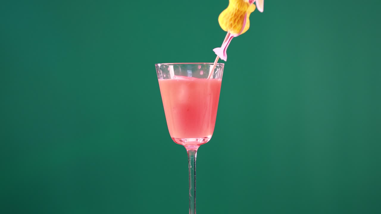 A pink cocktail in a glass with a decorative garnish against a green background. Bright lighting enhances the vibrant colors