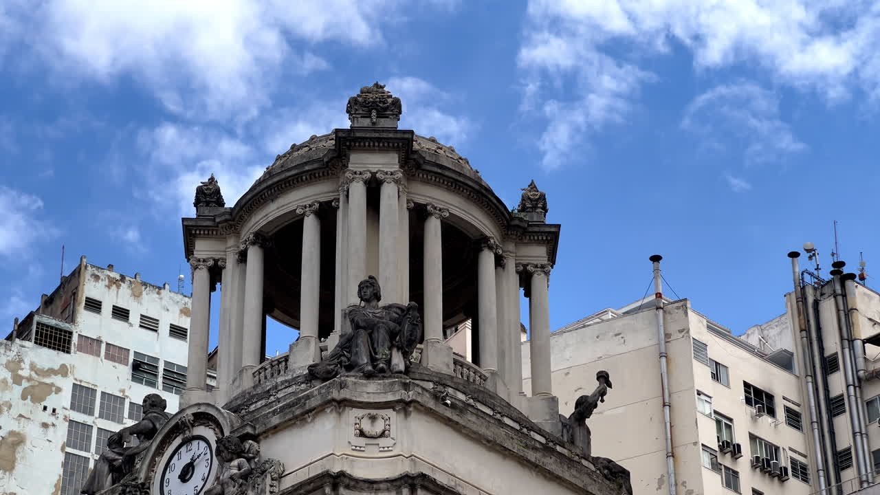 Close-up of ornate Neoclassical architecture in Rio de Janeiro's historic center. Features a detailed clock, dome, and classical statues set against a modern city background and blue sky