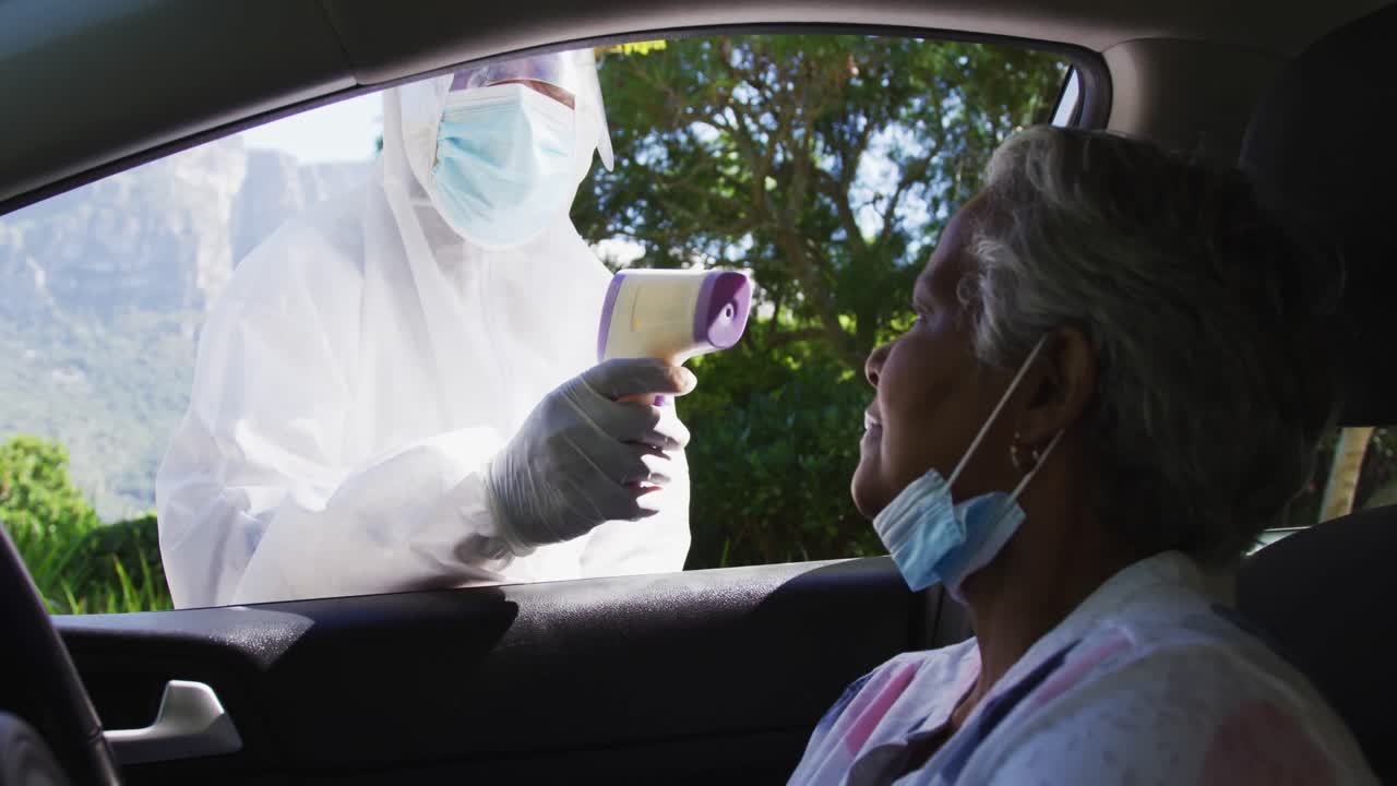 trabajador de la salud con ropa de protección midiendo la temperatura de una mujer mayor afroamericana en un coche