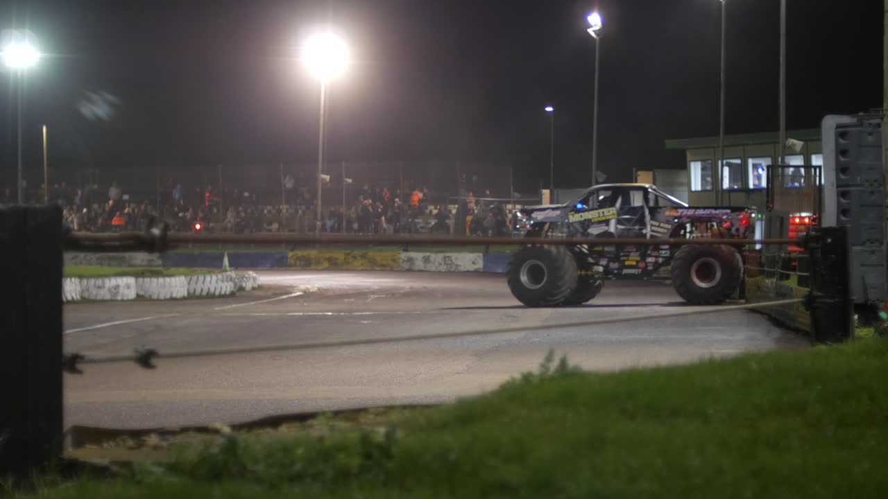 A massive monster truck enters the lit track, with its oversized tires and powerful build. Spectators in the stands watching as the vehicle prepares for an exciting stunt in the nighttime stadium