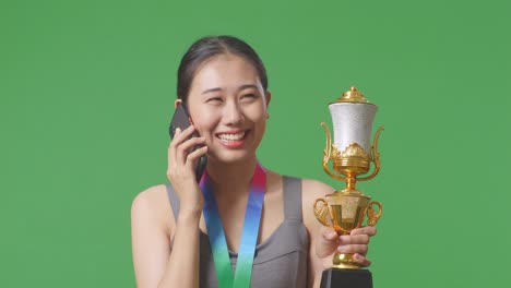 close up of asian woman with a gold medal holding a gold trophy and talking on smartphone on green screen background in the studio