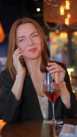 a cheerful woman is chatting on her phone and happily sipping a colorful cocktail at a vibrant bar setting