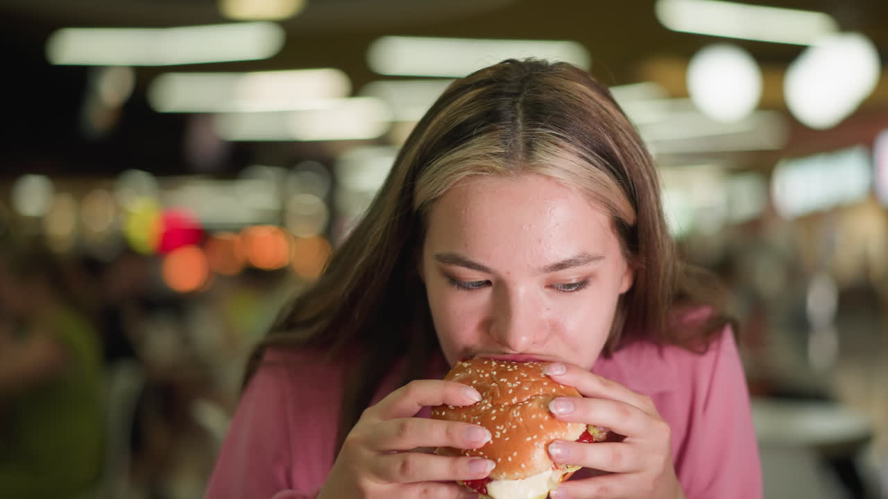 dama de vestido rosa toma un bocado de hamburguesa, mastica lentamente, y luego lo deja caer de nuevo en la bandeja mientras mira su mano abierta, el fondo presenta un efecto de luz bokeh borroso en un restaurante