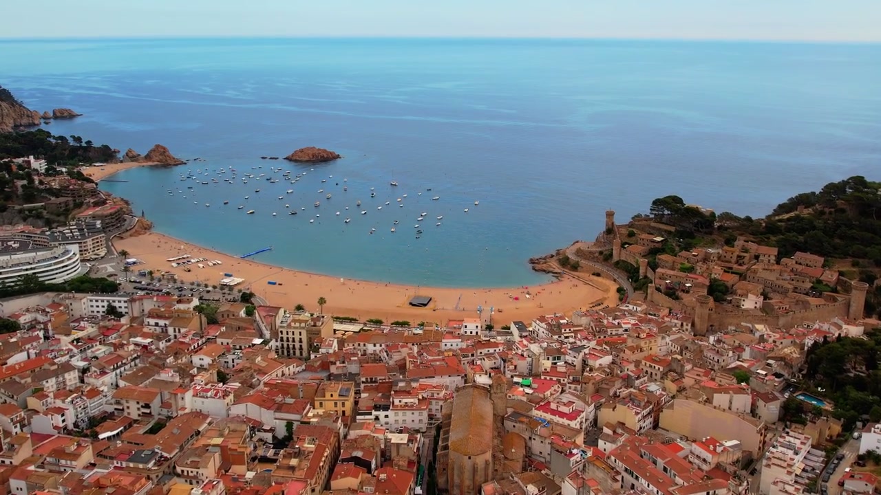 Eine Luftaufnahme von Tossa Del Mar in Spanien, die nach oben schwenkt und sich in die Stadt hineinbewegt, wobei die Bucht, Segelboote und die Küstenlinie in einer atemberaubenden Ansicht enthüllt werden.