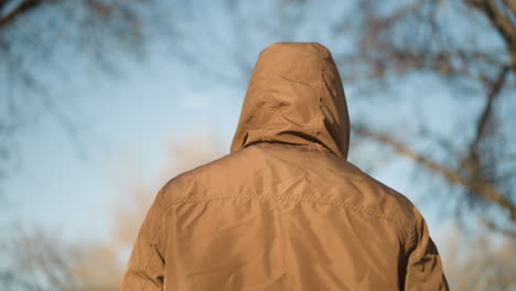 anonymous person walking outdoors in a brown hooded jacket against a blurred tree backdrop, capturing a sense of solitude and mystery