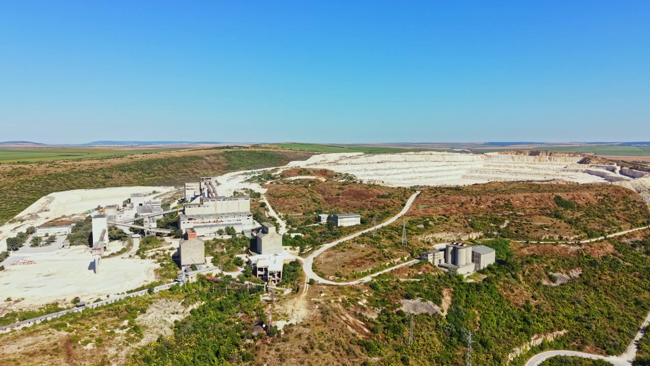 Pedreira mostrando máquinas, edifícios e terreno ao longe, com céus azuis claros e campos abertos