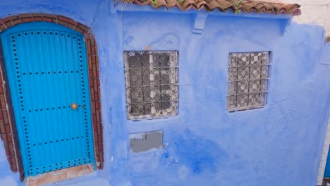 Vista-Panorámica-Izquierda-A-Través-De-Las-Paredes-Pintadas-De-Azul-Y-La-Puerta-Delantera-Del-Edificio-En-Chefchaouen,-Marruecos