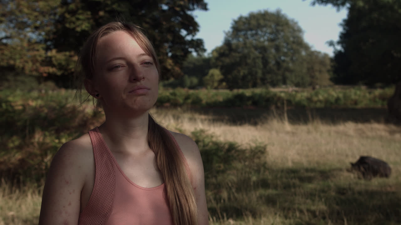 나무 아래에서 휴식을 취하고 공원의 풍경을 바라보며 태양 광선이 그녀의 얼굴에 떨어지고 있습니다.