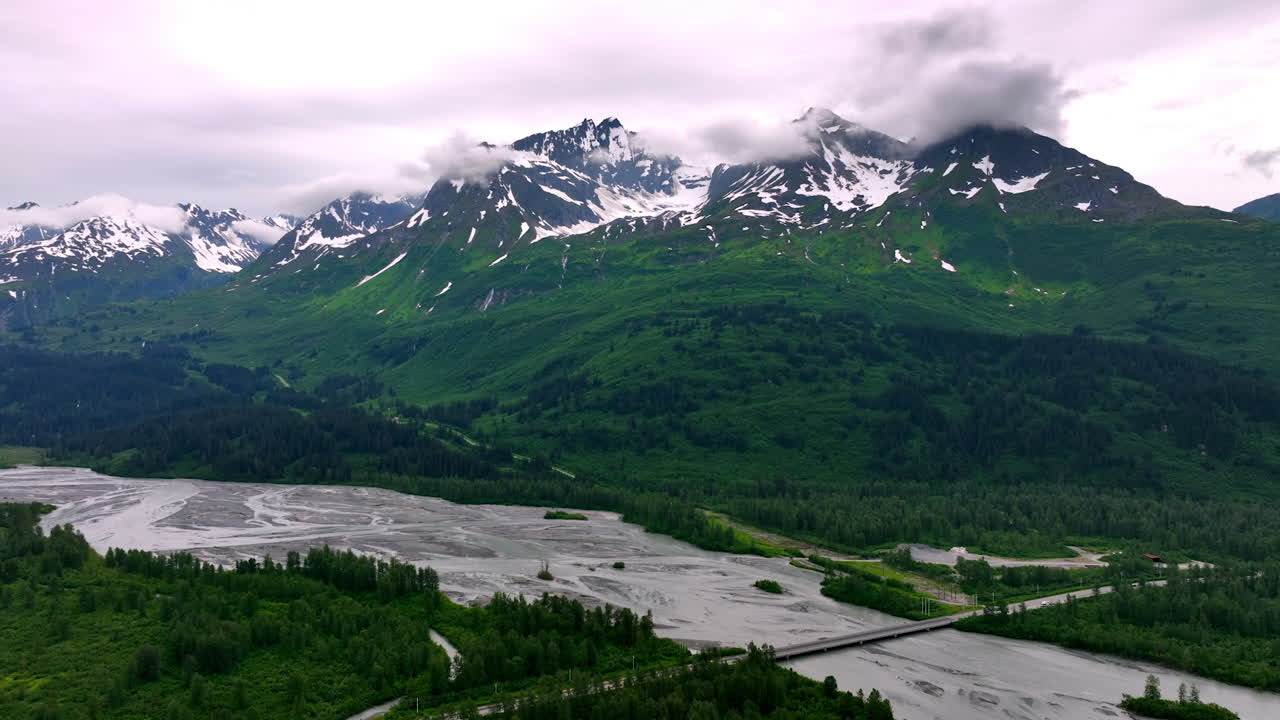 Glacial River and Forest Under Cloud-Covered Mountains. A wide vista features a light grey, braided glacial river delta and dense green forest in the foreground