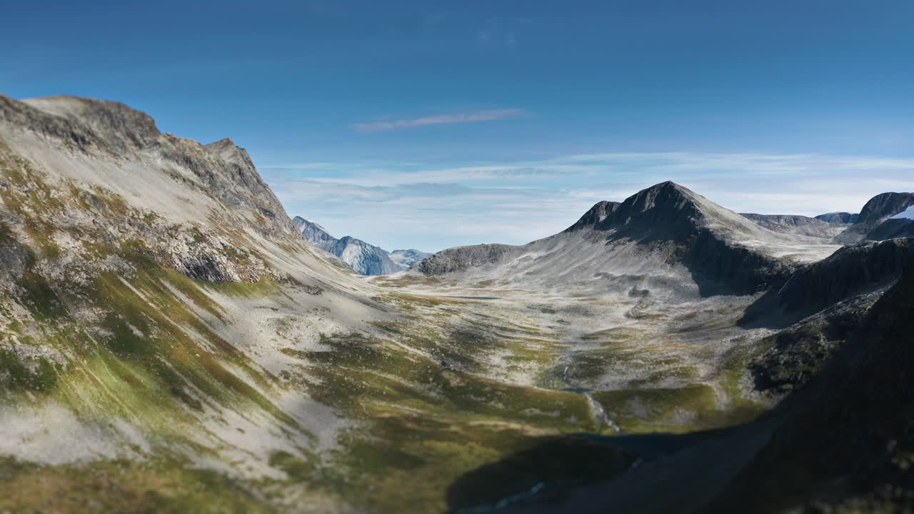 En vidstrakt fjell dal med ulendt, steinete terreng strekker seg under en lys blå himmel mens den endrer seg fra en grønn skog til et karrig, fremmedartet landskap. Flyfoto.