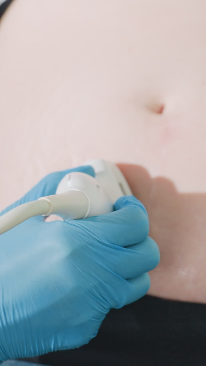 Primer plano de una mano enguantada sobre el abdomen durante una ecografía abdominal rutinaria alrededor del ombligo; ecografista ambulatorio aplicando gel suave y moviendo el transductor sobre la piel; guante de látex azul y cable en el encuadre.