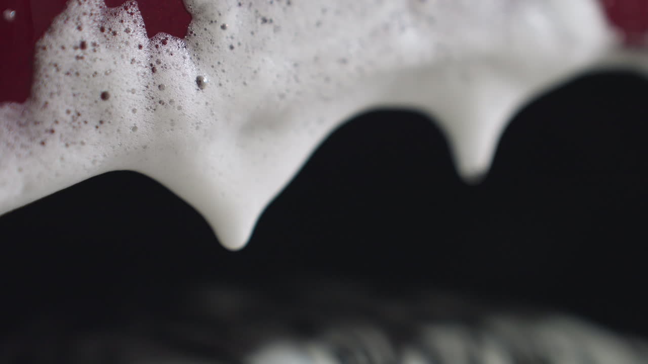 Extreme close up of soapy foam drop forming and dripping from car surface during washing process, creating dynamic view of liquid tension and water movement against dark blurred background