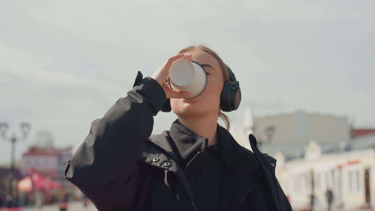 Giovane donna si gode una pausa caffè all'aperto, fiera di paese sullo sfondo, sorseggia dalla tazza con le cuffie, sguardo pensieroso e leggera brezza, cappotto e zaino, atmosfera da viaggio e rituale rilassato