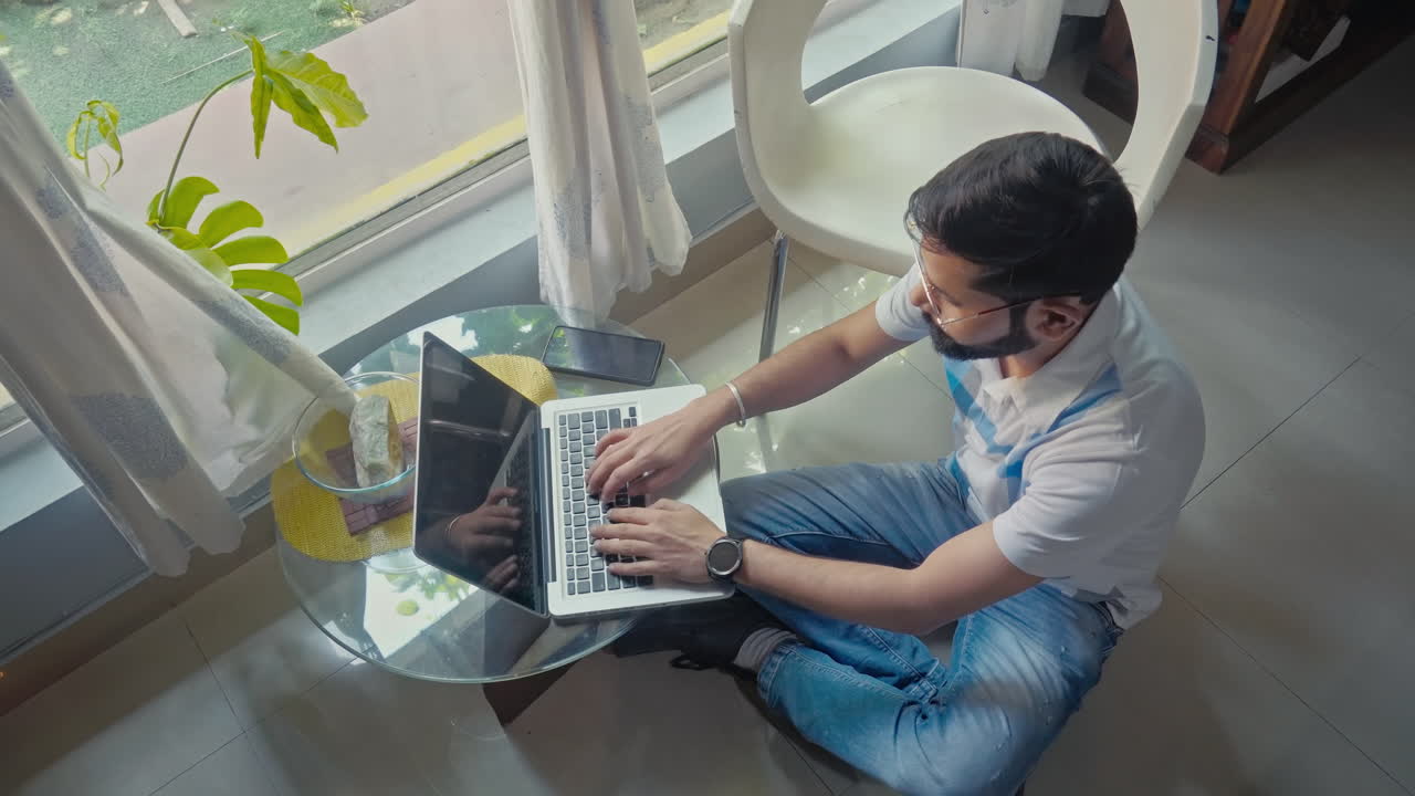 Laptop, wireless technology, internet. Top view of an Indian adult male using laptop while sitting on the floor near window side at home.