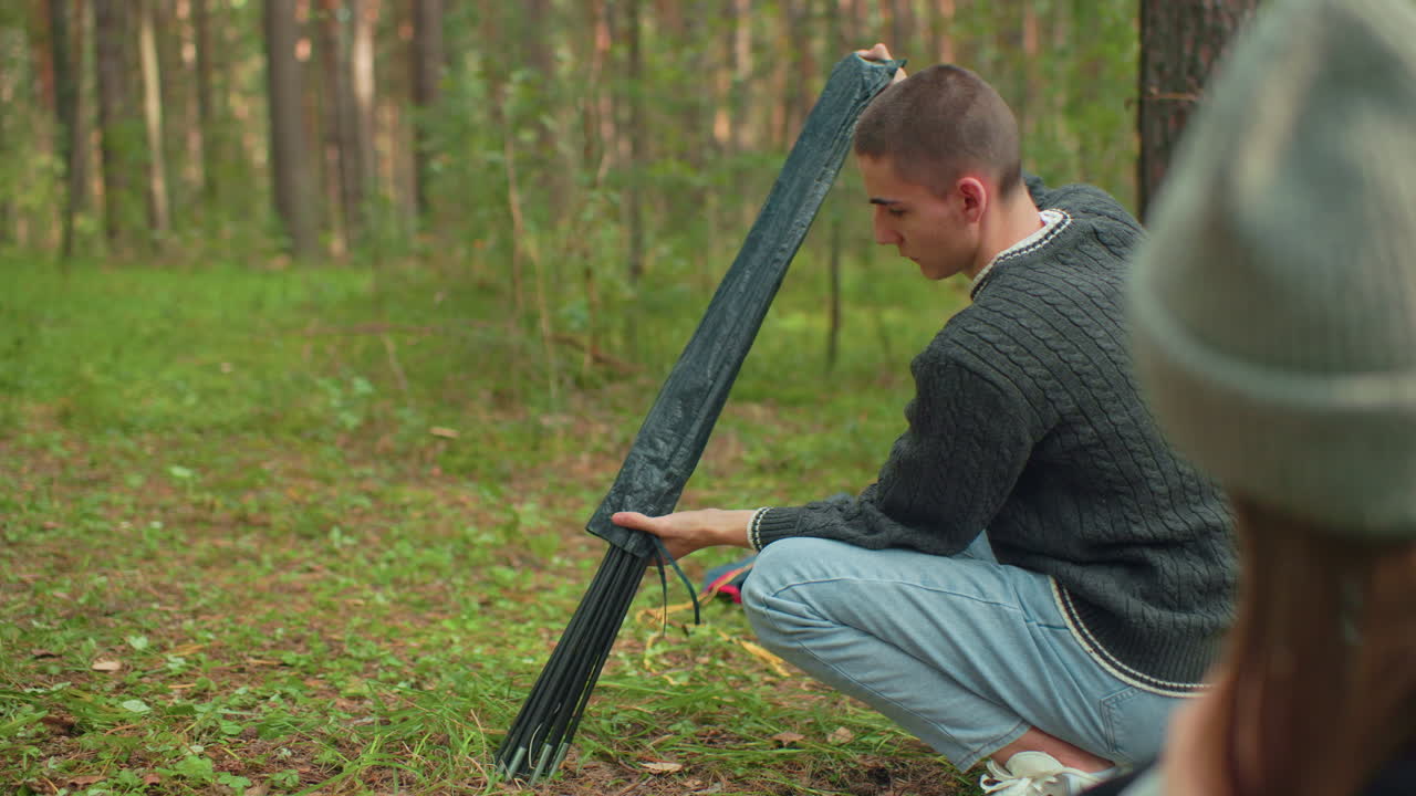 Close up of head view of woman in beanie observing man carefully remove tent pole from black cover while preparing to set up tent during peaceful forest camping activity on green grassy ground