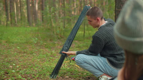 close up of head view of woman in beanie observing man carefully remove tent pole from black cover while preparing to set up tent during peaceful forest camping activity on green grassy ground