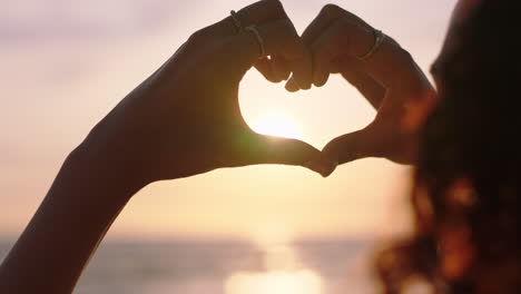 clsoe up woman hands making heart shape gesture holding beautiful sunset flare enjoying romantic vacation on beach