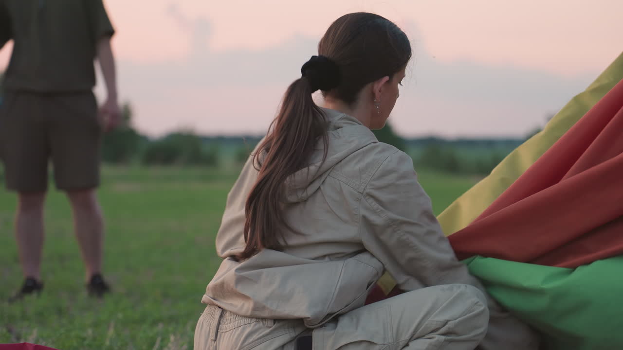 woman packing deflated colorful hot air balloon canopy into neat folds on grassy field while blurred couple hug under pastel sunset sky capturing end of flight routine and light breeze