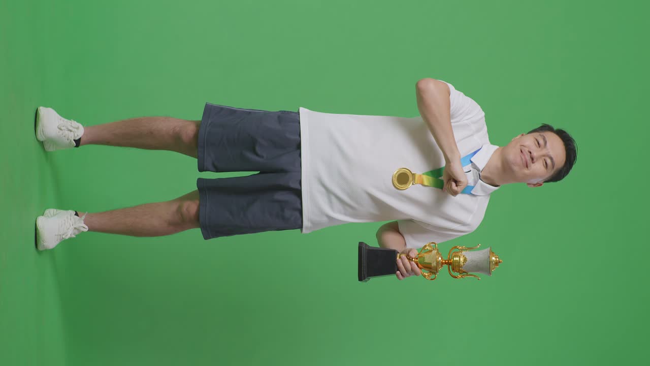 금메달을  아시아 남자의 몸은 카메라에 황금 트로피를 보여주며 미소 짓고 자신을 가리키며 스튜디오의 녹색 스크린 배경에서 첫 번째 우승자로 자랑스럽게 승리합니다.