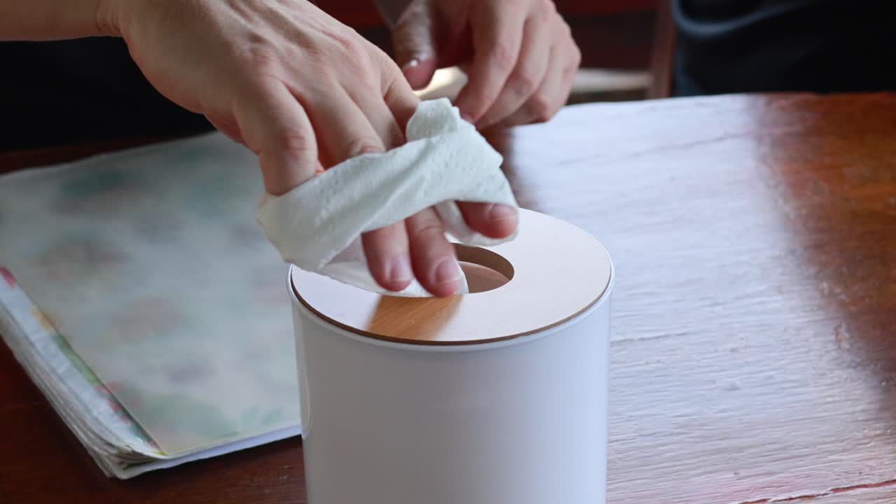 Hands pull tissue from a dispenser on a wooden table in natural light, showcasing a simple cleaning action
