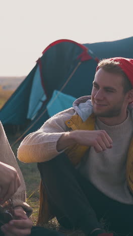 smiling young men talk resting at blue tent in camp on dry grass meadow in warm autumn evening close up