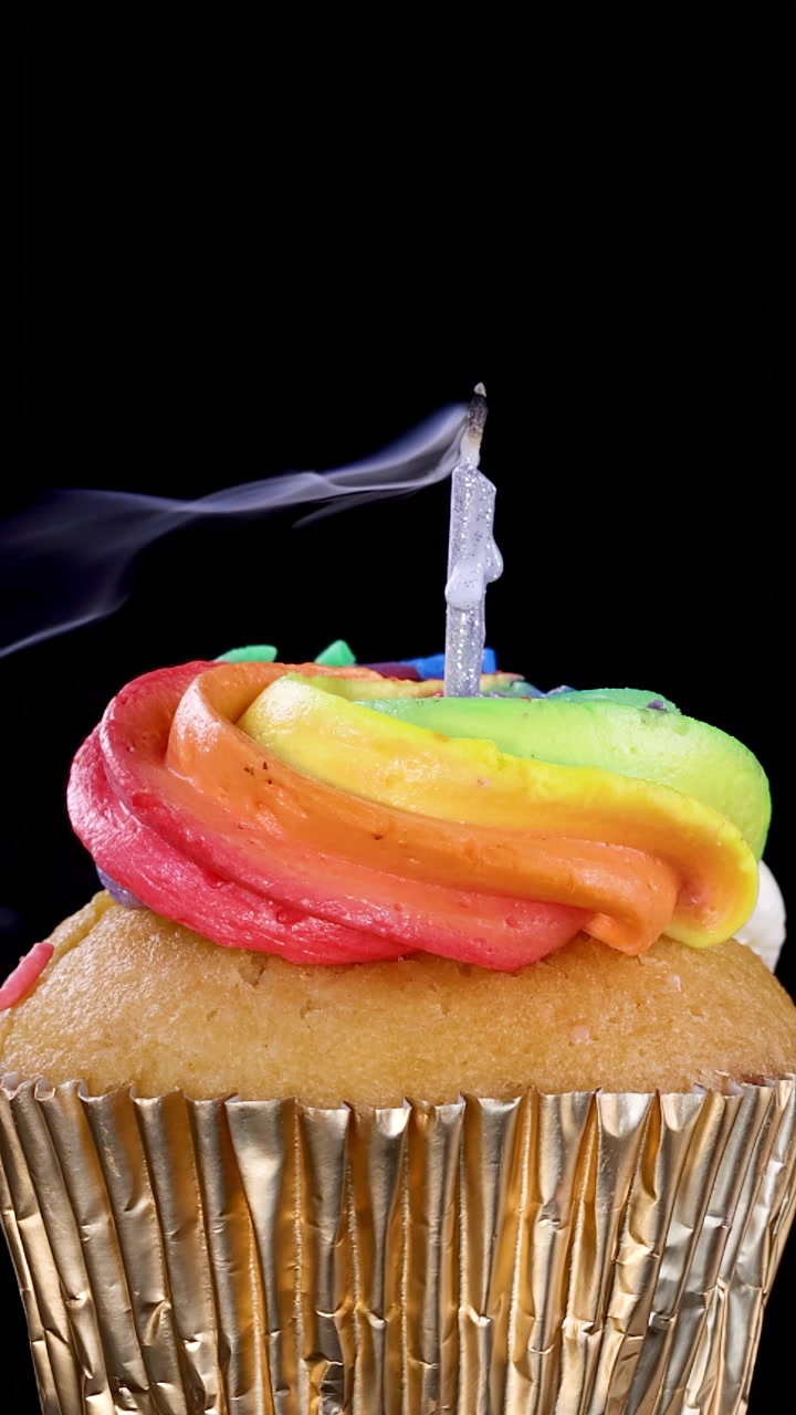 Un singolo cupcake con glassa arcobaleno e una candela appena spenta emette fumo a spirale su sfondo nero, ripreso in primo piano con illuminazione drammatica.